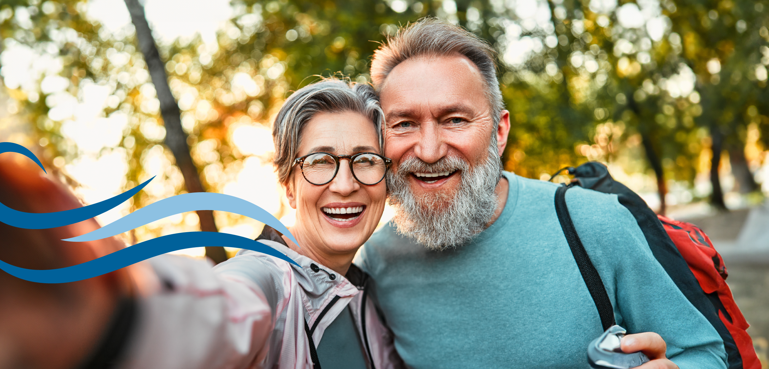 Woman in a pink jacket taking a selfie with a man in a blue shirt with a red backpack in front of trees. 